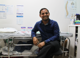 A health professional sits smiling in a clinic room in front of a patient bed and other medical supplies.