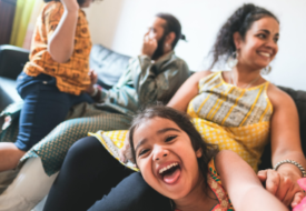 A family of four sits together on a couch laughing and playing
